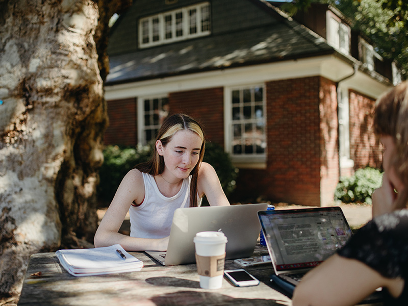 Reed students study in the shade of a tree next to a brick building